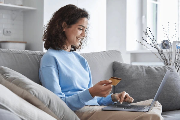 Woman sitting on a couch using a laptop and holding a credit card in a home setting.