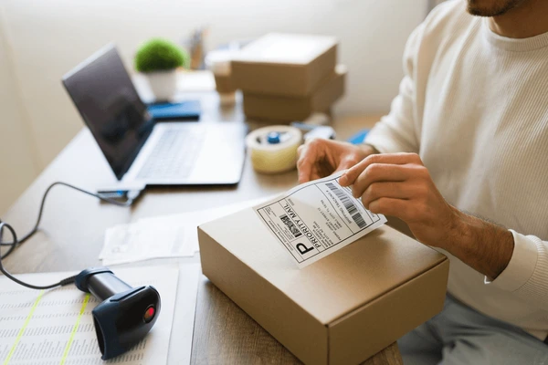 Person preparing a package with a label, sitting at a desk with a laptop and scanner.